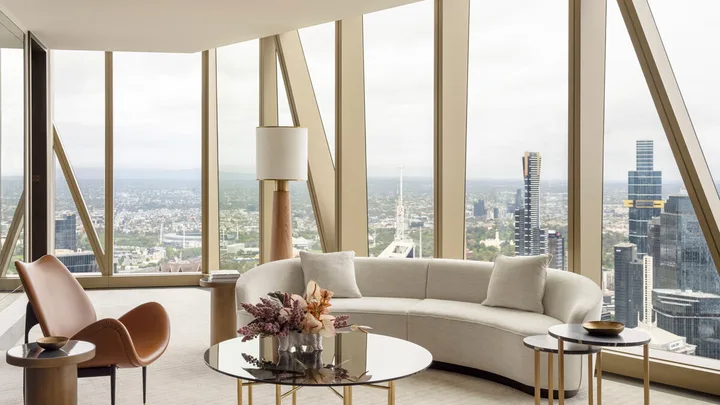 Modern living room with large windows, city view, beige curved sofa, glass coffee tables, and a brown chair.