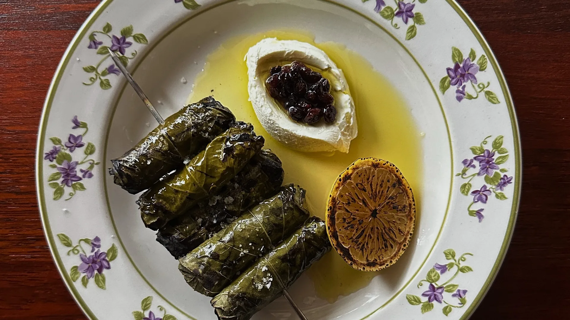 An overhead shot of a vintage plate with a skewer of four folmades, olive oil, dip and a charred lemon half.