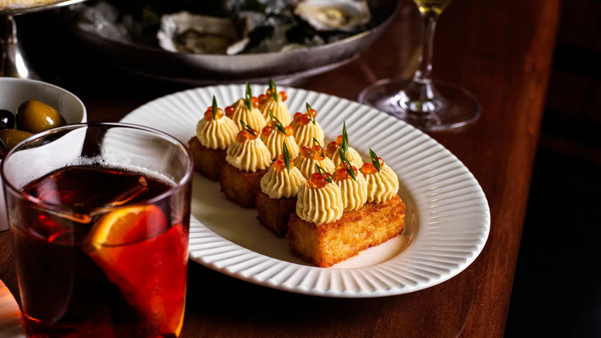 A close-up shot of golden, crisp potato hash topped with beautifully whipped cod roe, roe pearls and chives, alongside a Negroni, for happy hour at Bistro George