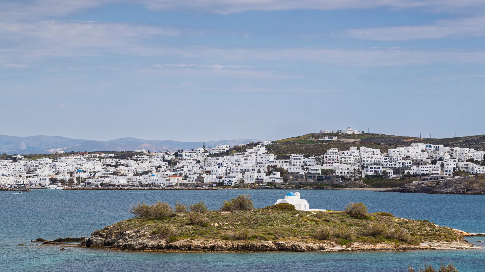 View of a small island with a traditional white and blue Greek chapel and the village of Naousa (Naoussa) on Paros island in Greece on a sunny day.
