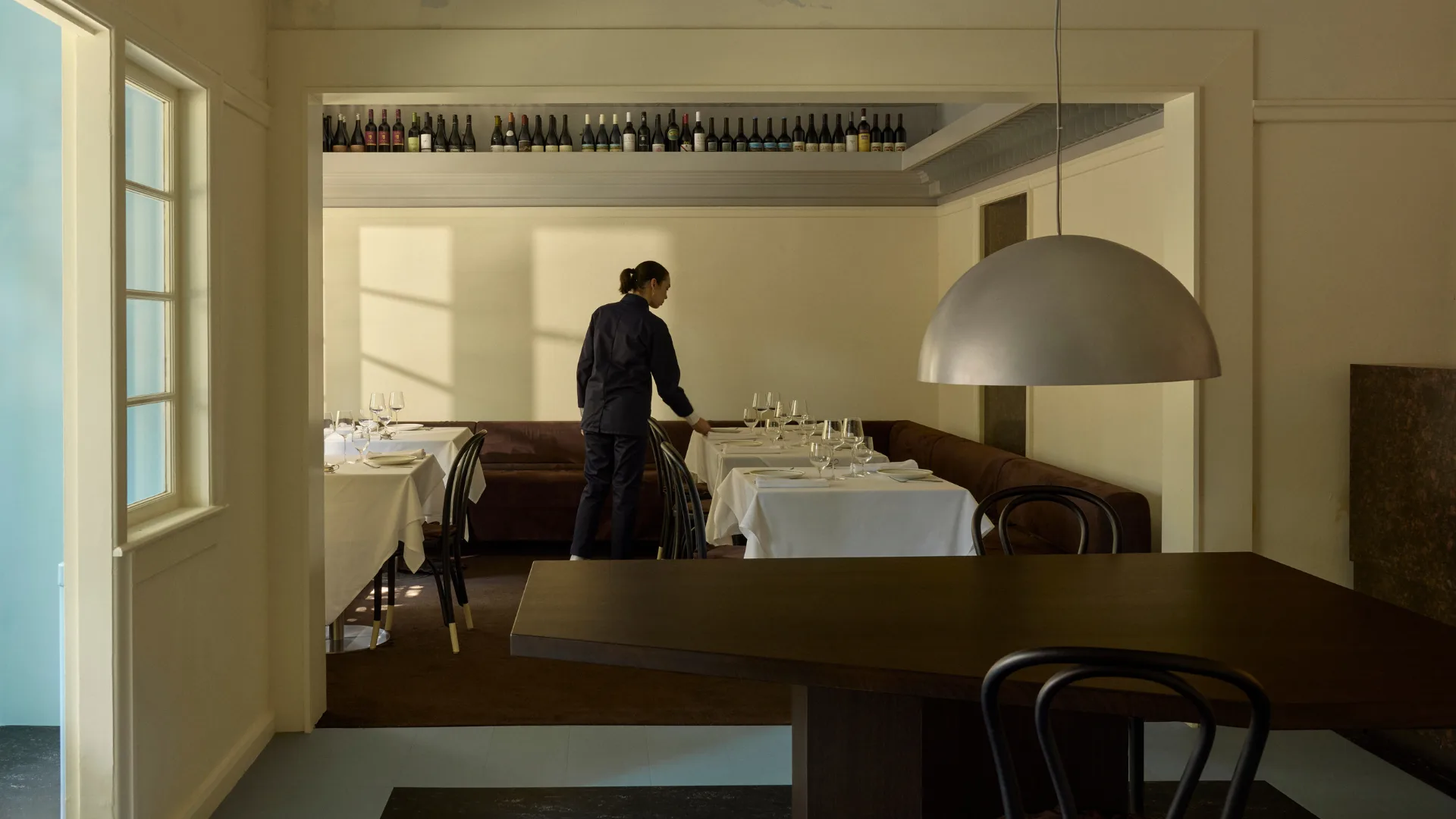 A waiter sets a table in South Brisbane restaurant Marlowe, with wine bottles on display and a statement metal light shade