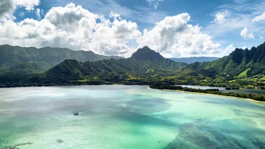 Aerial view of a coastal area on Oahu, Hawaii. Turquoise waters with visible coral reefs, forested land and steep green mountains.