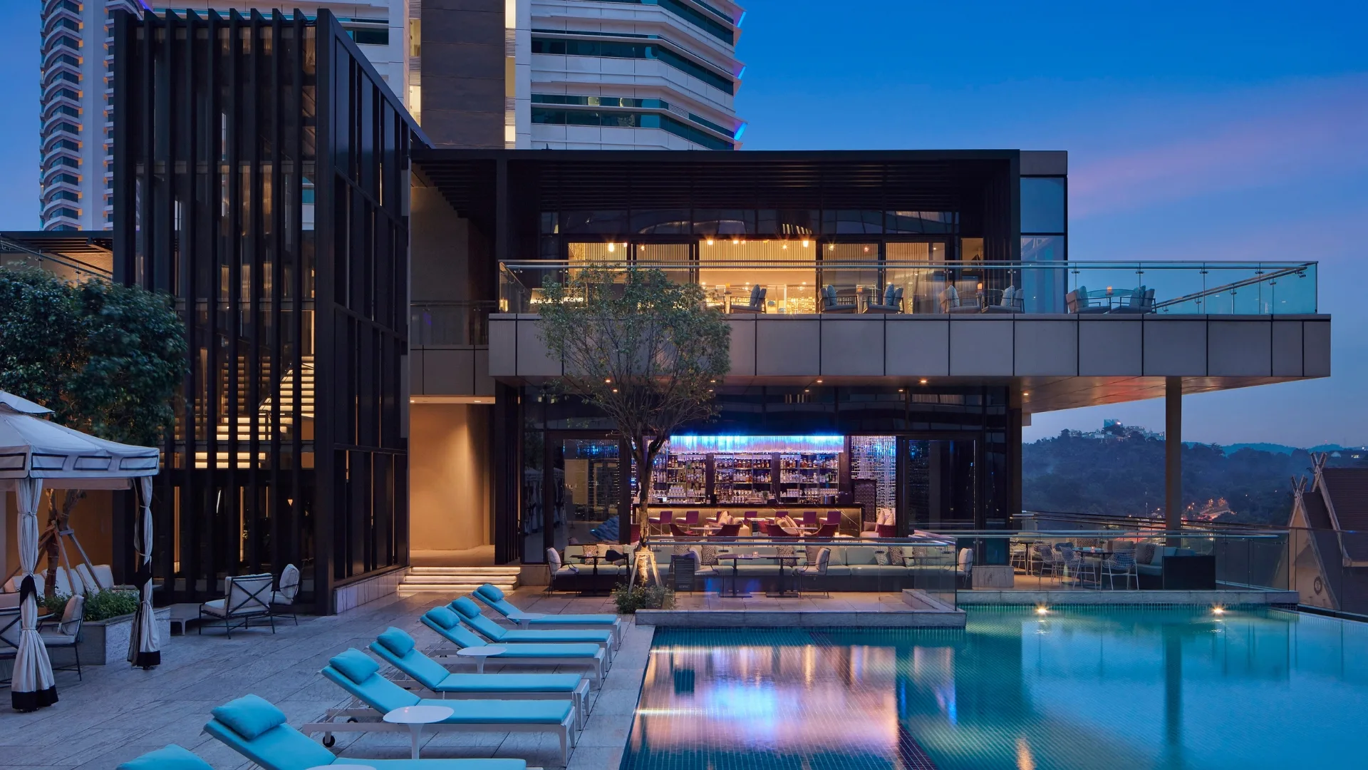 A photo of The St. Regis in Kuala Lumpur at dusk, featuring an open-air deck with pool chairs, a large pool and indoor bar
