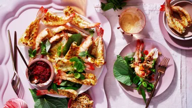 Butterflied barbecued prawns on a pink plate and tablecloth, accompanied by mint leaves and a small bowl of sambal