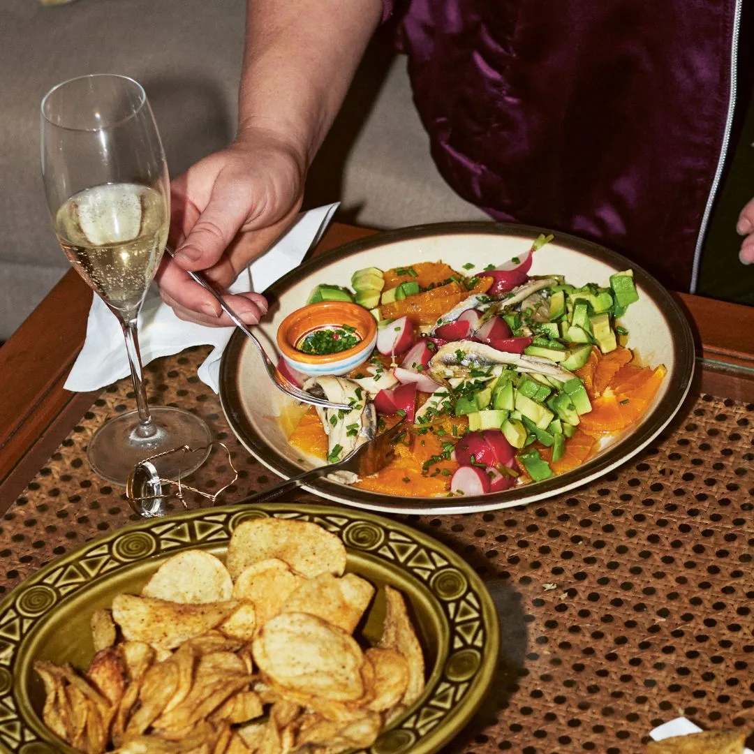friendly anchovies salad in a bowl with oranges, white anchovies, avocado and radishes, alongside a glass of champagne and bowl of potato chips.