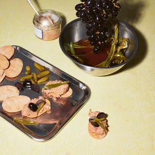 a silver tray of crackers topped with with duck rillettes, cornichons and pickled black grapes, with an additional bowl of black grapes to the side
