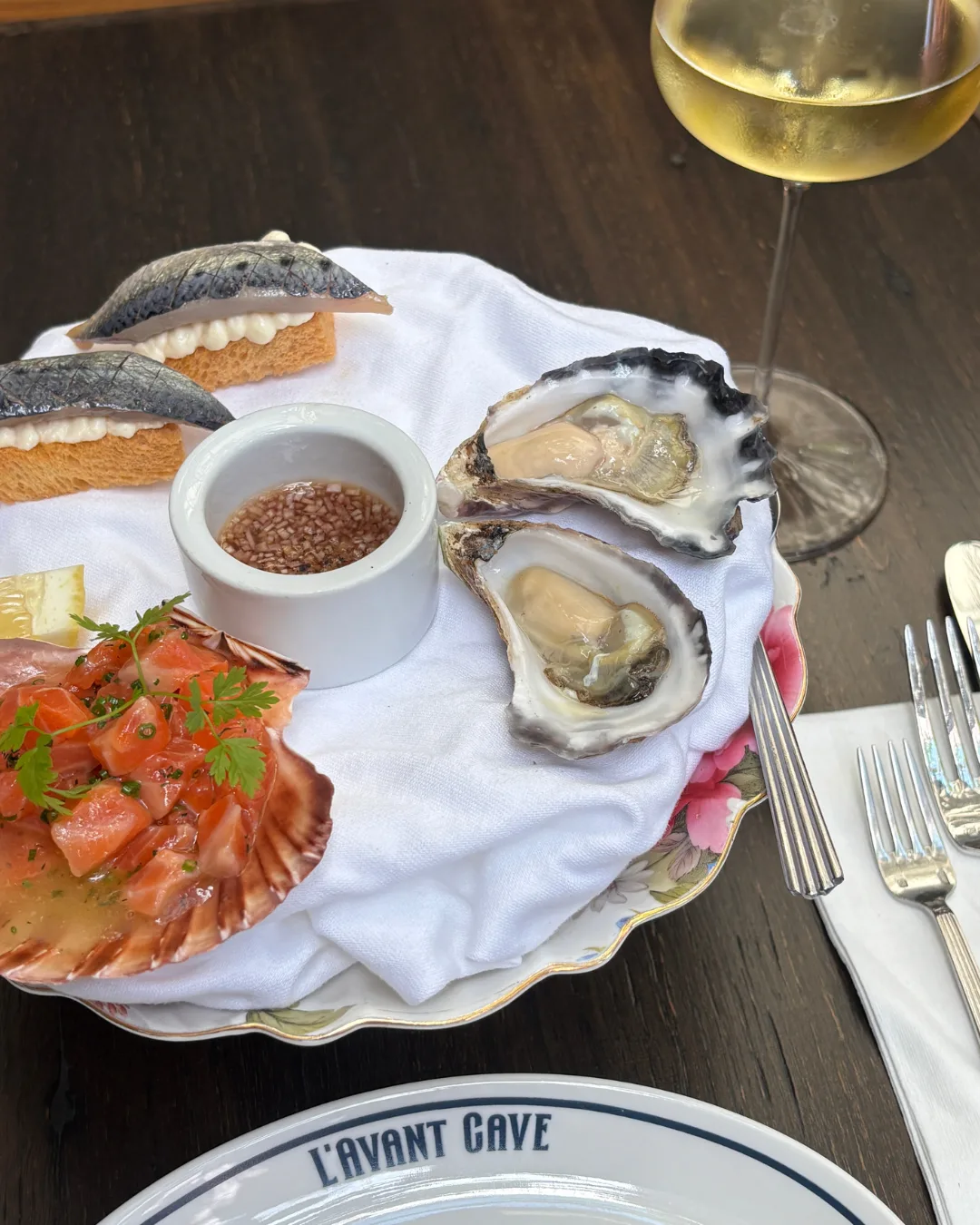 A platter of oysters, trout crudo served in a scallop shell, and toasts topped with sardine, next to a glass of white wine.