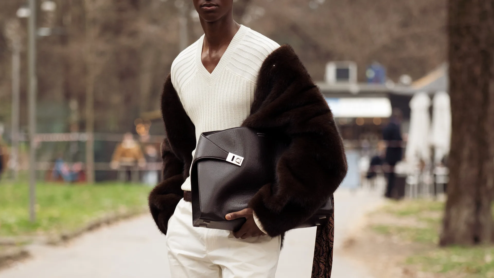 a man wearing a white ribbed V-neck knit sweater layered under a dark brown fur stole draped over the shoulders, paired with white straight-leg trousers, carrying a dark leather clutch bag