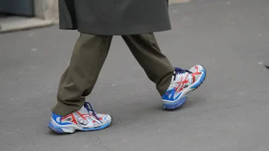 Left image a dark gray wool coat hem, green olive tailored trousers, white gray mesh and leather sneakers shoes with red overlays, blue accents, blue laces, and blue rubber soles, outside Prada, during Milan Fashion Week