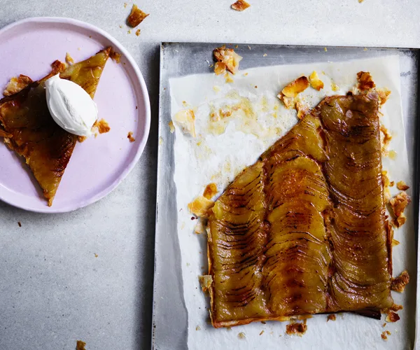 A flat apple tart, with the top-left corner sliced off, on baking paper and a metal tray.