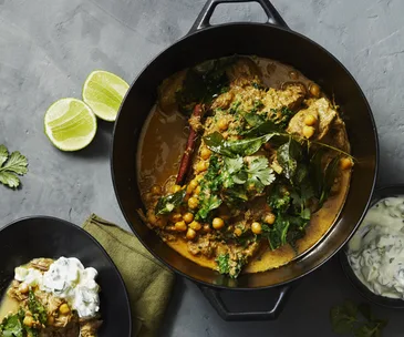 Overhead shot of a last cast-iron pot of lamb and chickpea curry, studded with cinnamon quills and garnished with coriander. A bowl of yoghurt, and sliced lime cheeks, sit to the side.