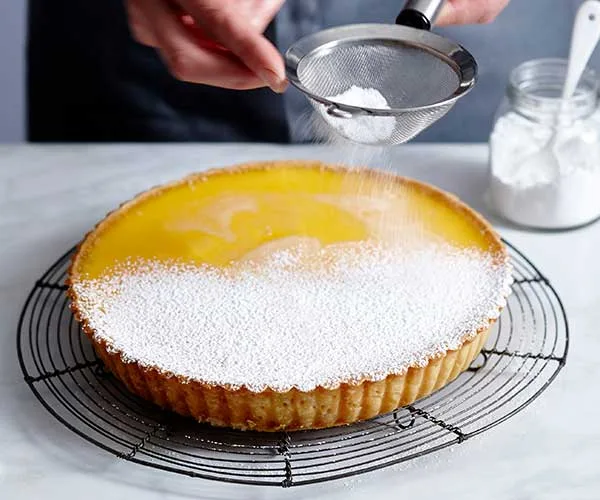 A whole lemon tart on a black wire rack being dusted with icing sugar.