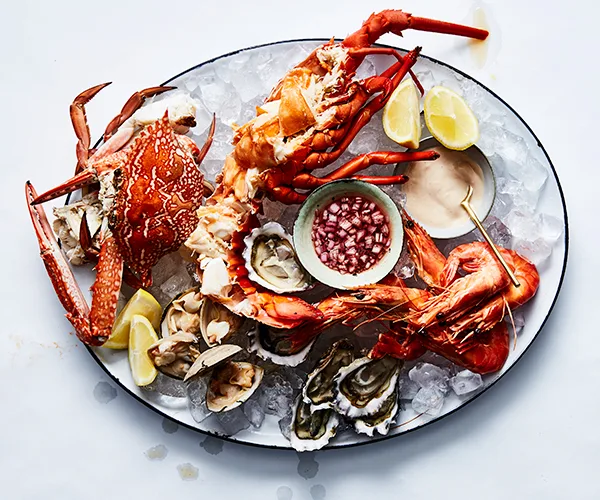 Over the top shot of a large white oval platter laden with seafood: a whole crab, a whole lobster, cooked prawns, clams, and shucked oysters.