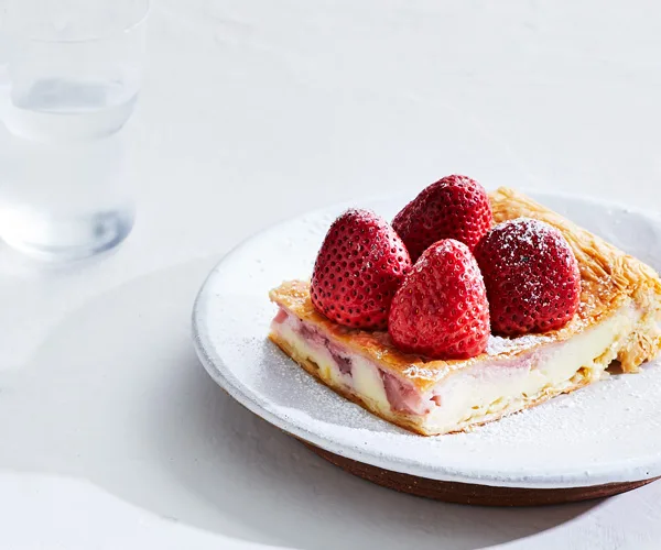 A square slice of galaktoboureko, topped with four strawberries, on a white plate.