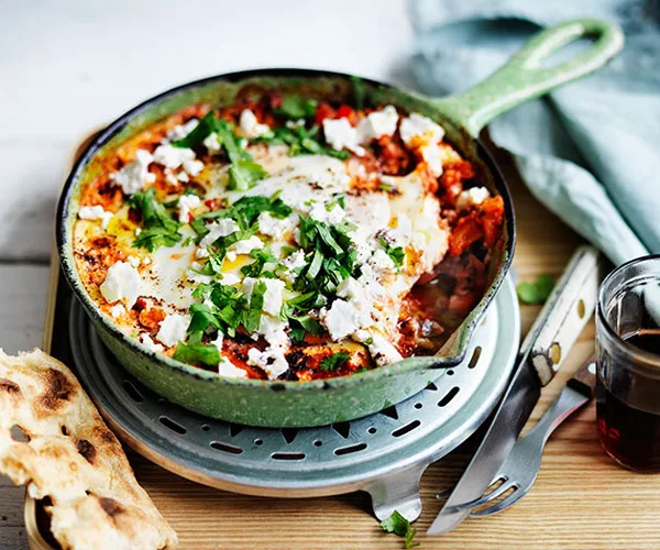 A green frying pan holding a mixture of fried eggs and tomato, topped with crumbled feta and chopped green herbs.
