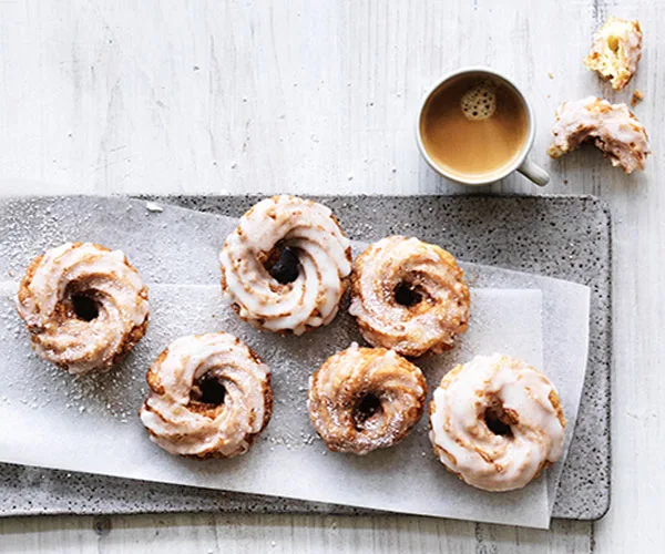 Five round glazed doughnuts with swirled ridges, on a grey rectangular plate, with a cup of tea in the top right corner.