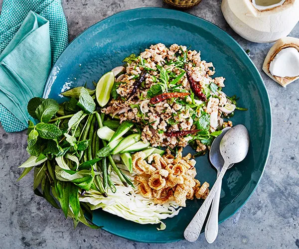 A blue plate with a salad of minced chicken and chillies, a mound of green herbs, shredded white cabbage, and roasted peanuts, with a silver fork and spoon.