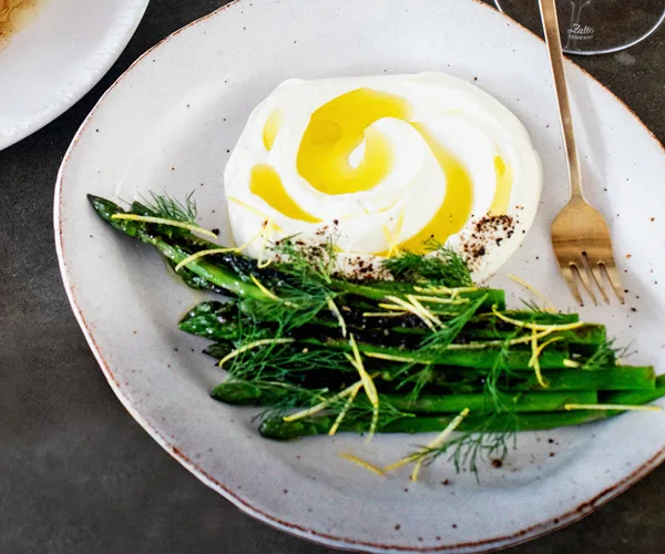 Asparagus spears, sprinkled with lemon zest and dill, with a mound of labne swirled through with olive oil, on an off-white plate with a gold fork.