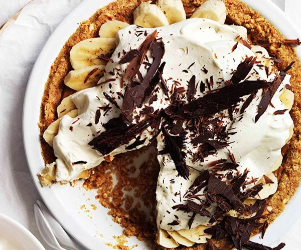 Pie with a golden, crumbly base, topped with sliced bananas, cream and chocolate shavings, with a slice taken out of the pie.