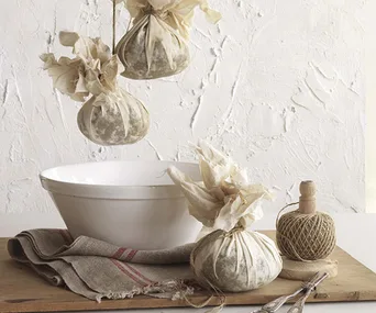 An arrangement displaying two hanging boiled Christmas puddings, wrapped in calico and tied with string, and a table arranged with a white pudding bowl, a grey tea towel, kitchen string, scissors and a wrapped Christmas pudding