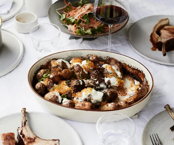 Photo of white baking dish filled with creamy potato bake surrounded by other plates and a glass of red wine