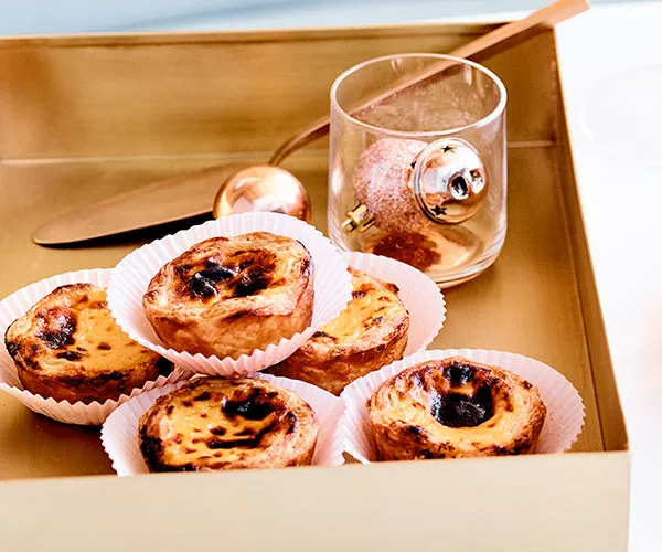 Golden square tin holding hand-sized custard tarts with burnished tops, each in their own white paper casing. There's also a glass holding a rose-pink speckled bauble, which is kind of weird, but we'll take it.