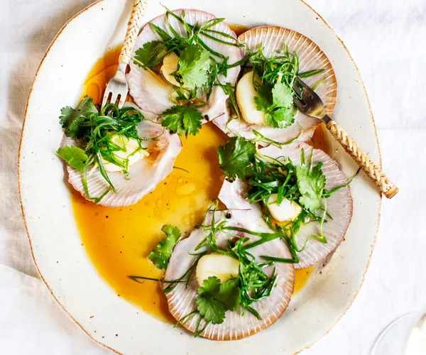 Five scallops in a half-shell topped with herbs and a soy dressing, on an off-white plate, on a white marble background.