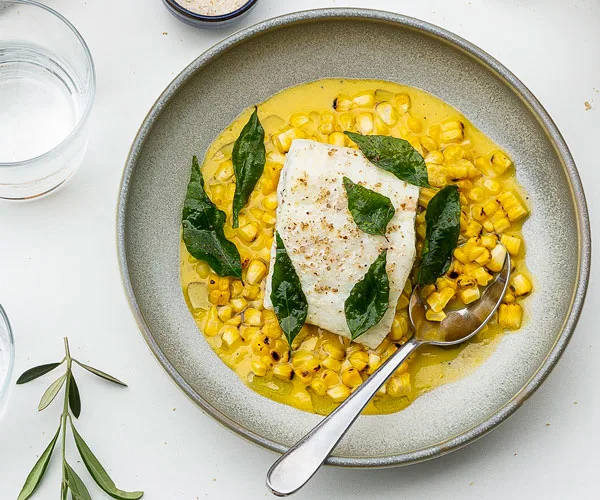 Bird's eye view image of grey ceramic bowl displaying yellow corn custard, white fish fillet and seven fried curry leaves, plus a silver spoon