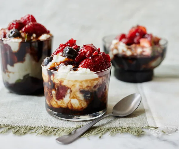 Three glasses, one in the foreground, two in the back, holding a tumble of mixed berries, goat's curd and balsamic vinegar. A spoon lies in the foreground.