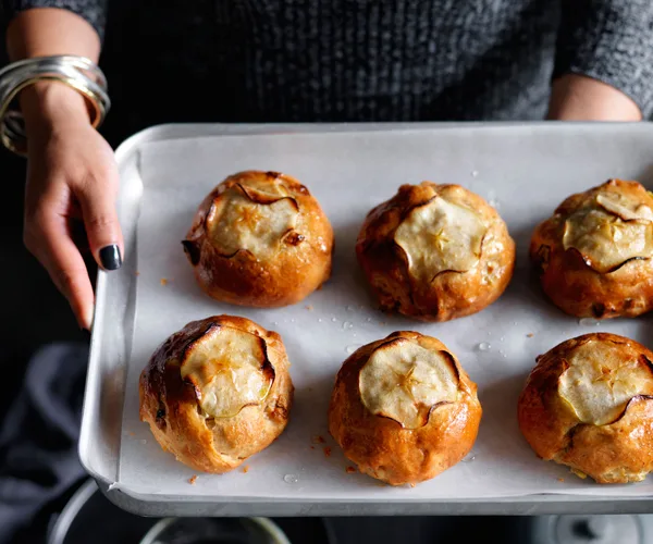 Six round apple buns on baking paper on a metal tray, held by a woman's hands painted with black nail polish