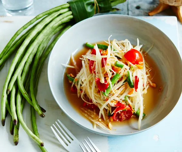 White bowl holding a salad of shredded green papaya, chopped green beans and halved cherry tomatoes. A bunch of snake beans is placed to the side.