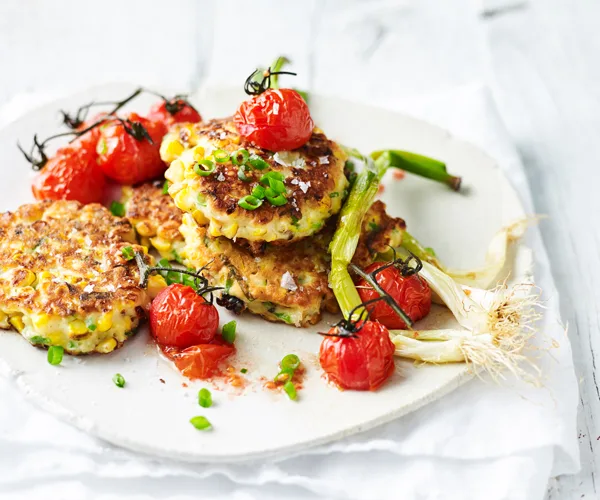 A white plate with fried corn fritters and roasted cherry tomatoes, garnished with chopped and whole shallots.