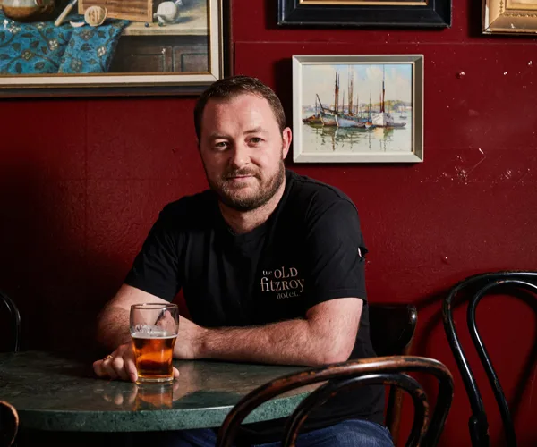 Chef Nik Hill sitting at a round table, with a beer in hand, with a red wall in the background.