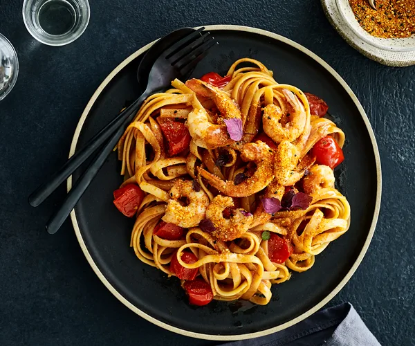 A black plate holding fettuccine, prawns and chunks of tomato, with a black fork and spoon to the side.