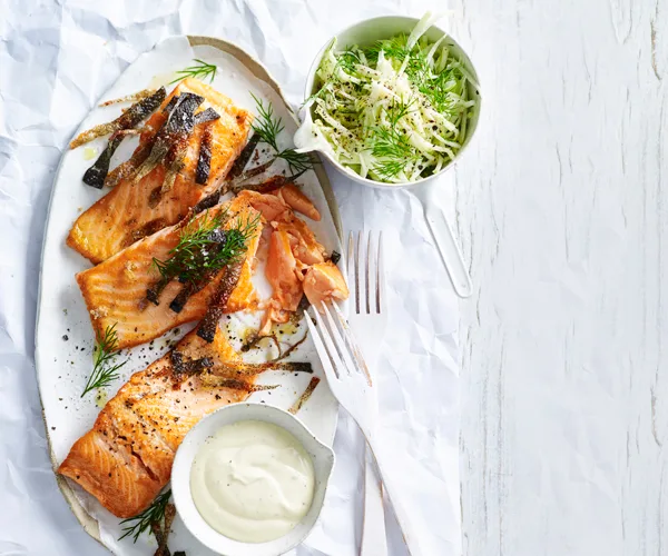 Three portions of ocean trout topped with strips of crisp fish and dill, with a bowl of yoghurt, on a white oval platter. A bowl of shredded kohlrabi is position at the top-right.