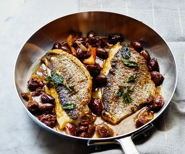 Two sides of white fish, skin side up, surrounded by black olives, in a metal fry pan, on a grey tea towel.