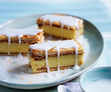 Three pieces of vanilla slice (custard sandwiched in pastry), drizzled with icing, on a blue and white plate.