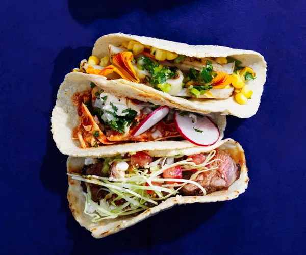 Bird's eye shot of three tacos, filled with calamari and corn, spiced chicken and radish, and beef, on a dark blue background.