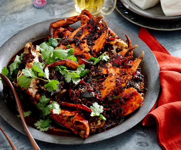 A grey plate holding cooked crab pieces covered in a black pepper sauce and garnished with coriander. A red napkin is arranged on the side.