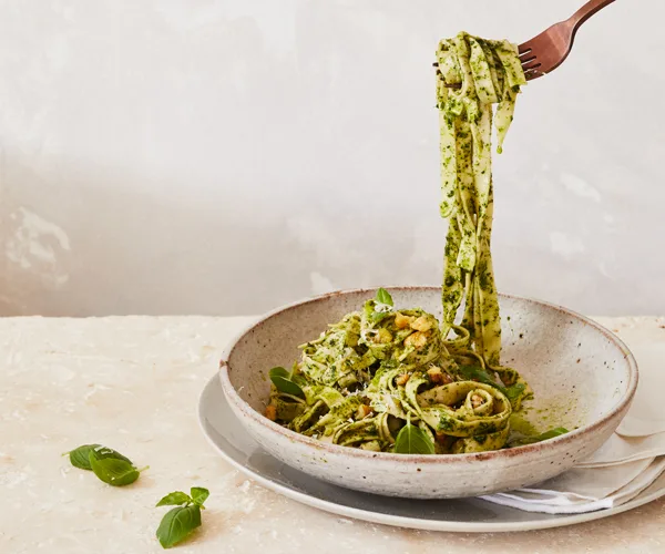 A raised fork dangling strands of long pasta coated in a green herb sauce, from a brown-white bowl.