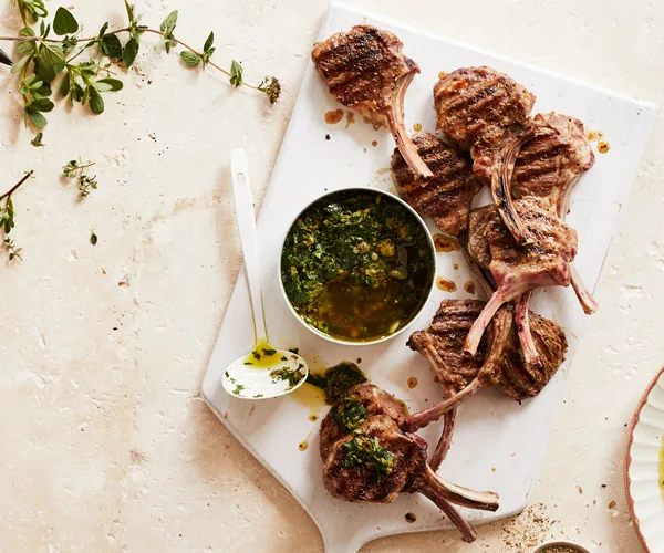 A white chopping board with grilled lamb cutlets, a bowl of herb sauce in olive oil, and a white ladle.