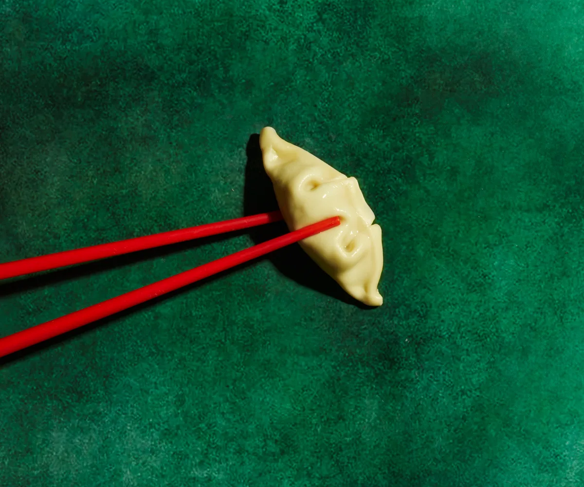 A single dumpling, grasped in a pair of red chopstick, on a dark green background.