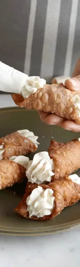 Person filling cannoli shells with cream on a plate, surrounded by preparation tools on a marble counter.
