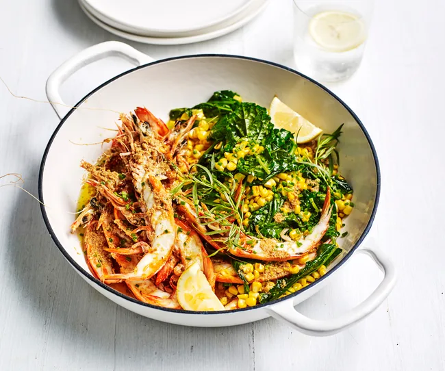 Grilled prawns with corn and greens in a white dish, garnished with lemon wedges and fresh herbs.