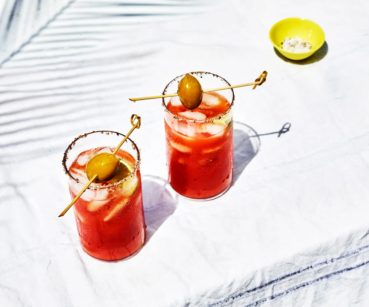 Two clear glasses filled with a red Bloody Mary liquid, topped with green olives on a skewer.