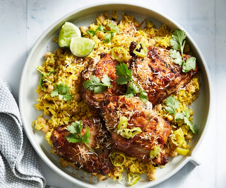 Close-up of a plate of roast chicken thighs on a bed of yellow pilaf, garnished with coriander leaves and lime wedges.