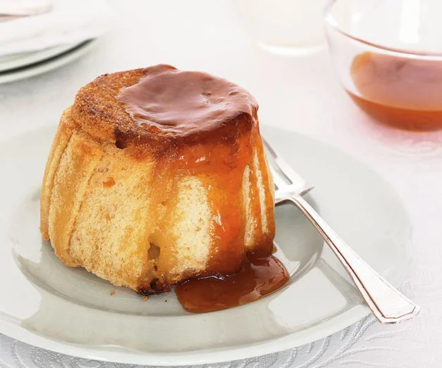 A steamed round apple pudding, drizzled with warm apricot jam, on a round plate with a silver fork on the right.