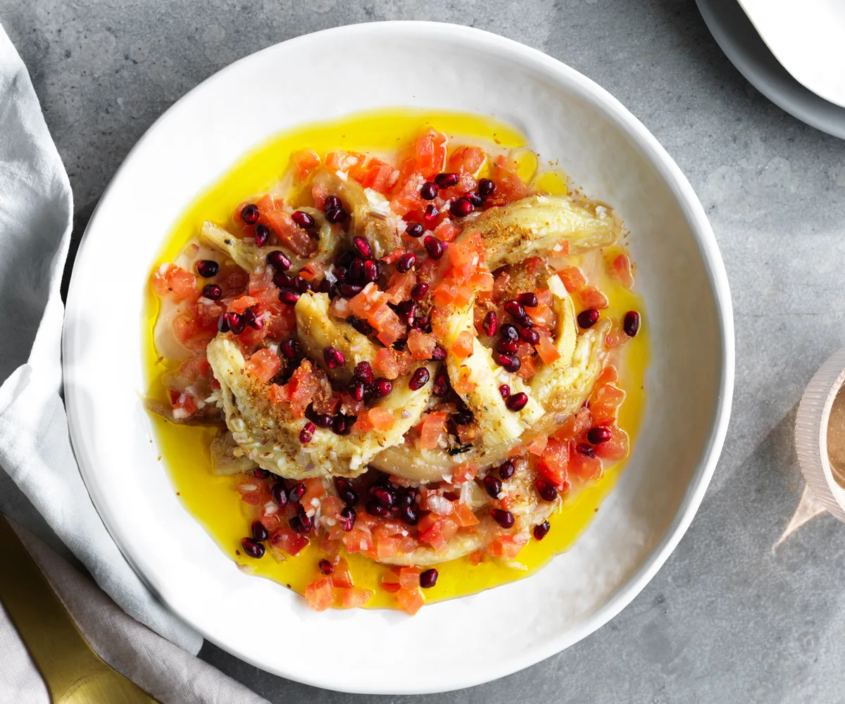 Overhead shot of grilled eggplant slices, finely chopped tomato, pomegranate seeds and olive oil on a round white plate.