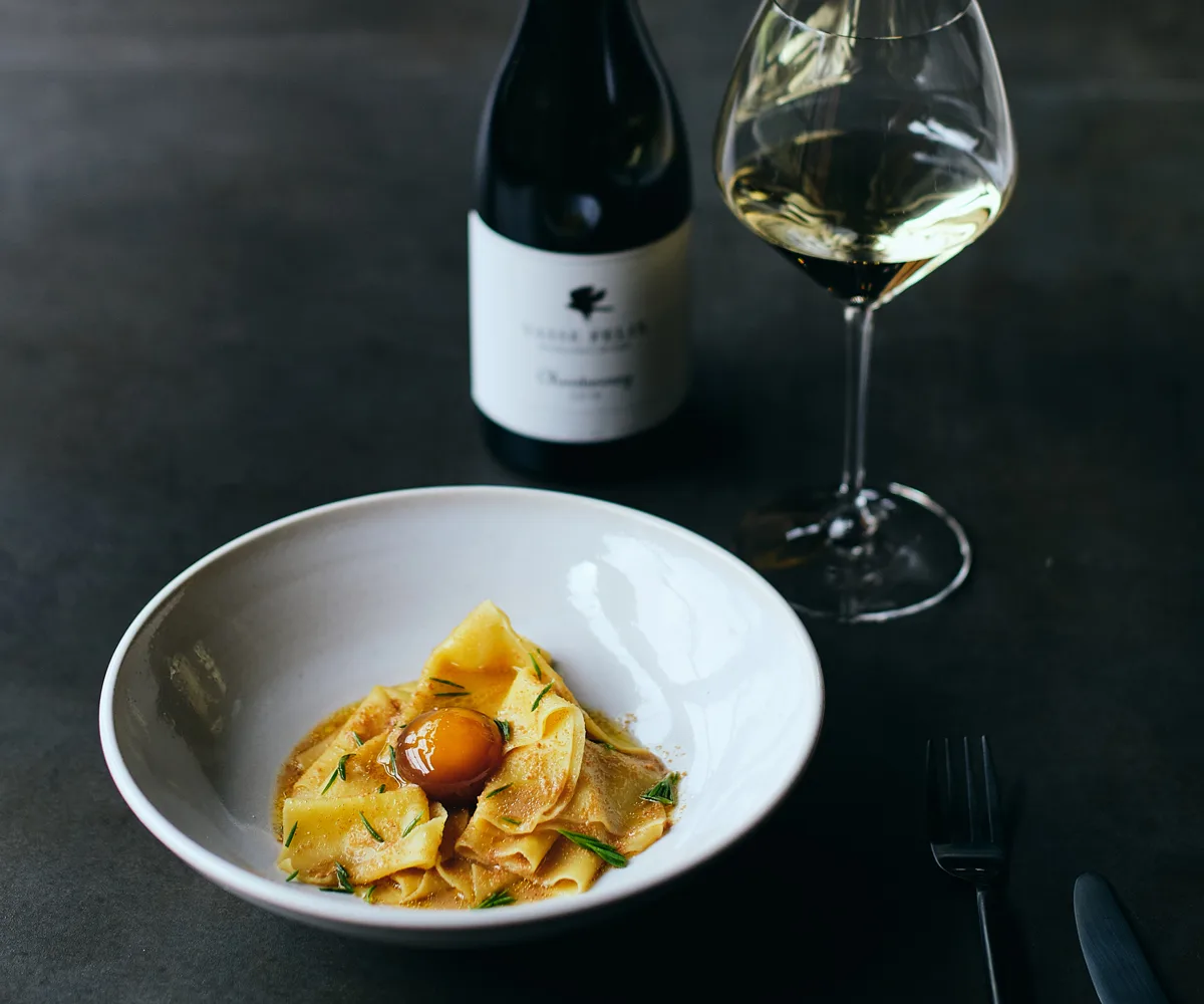A white dish with wide ribbons of pasta, a wine glass with white wine, and a wine bottle.