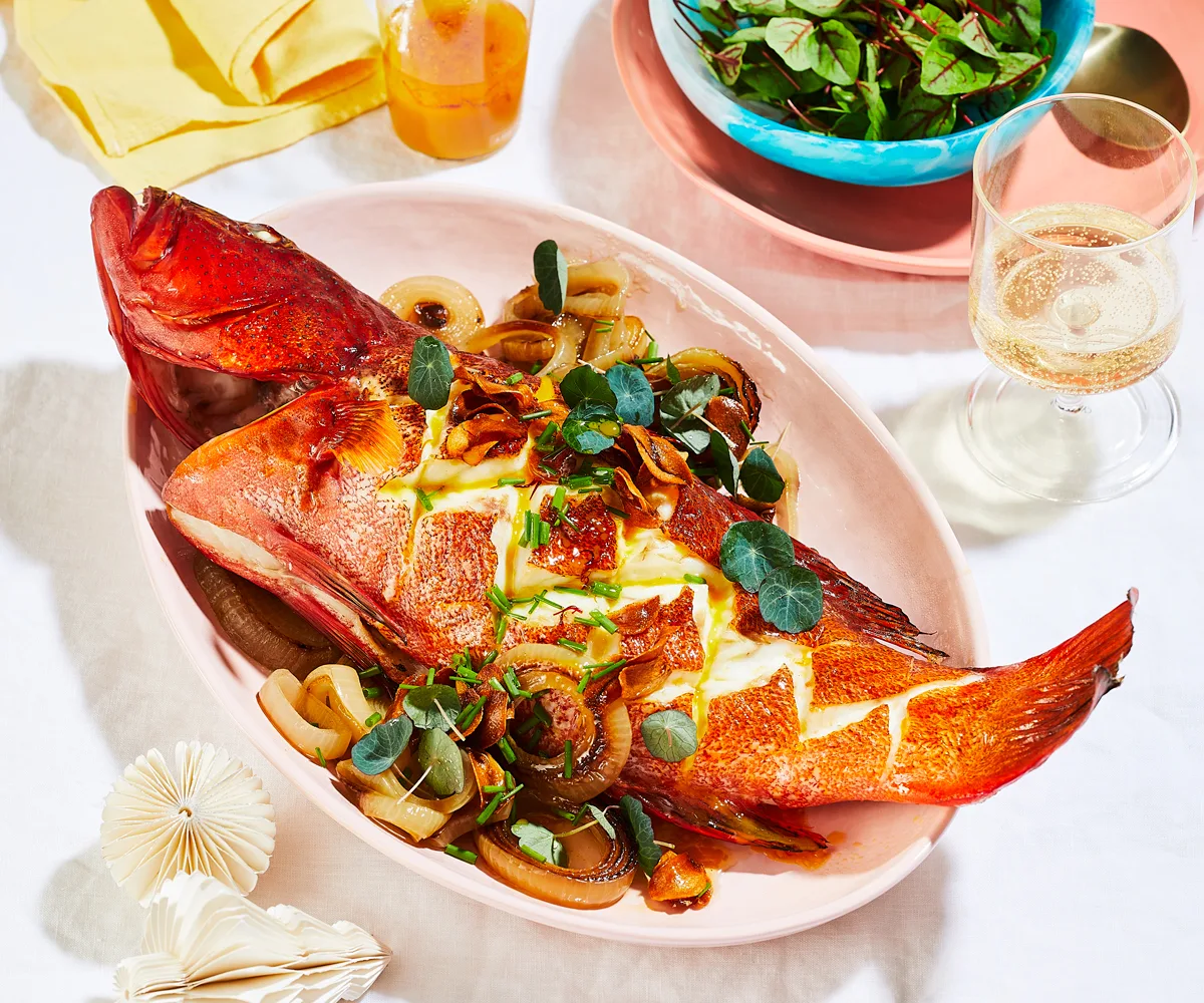 A baked trout on a pink oval platter, garnished with roasted onions